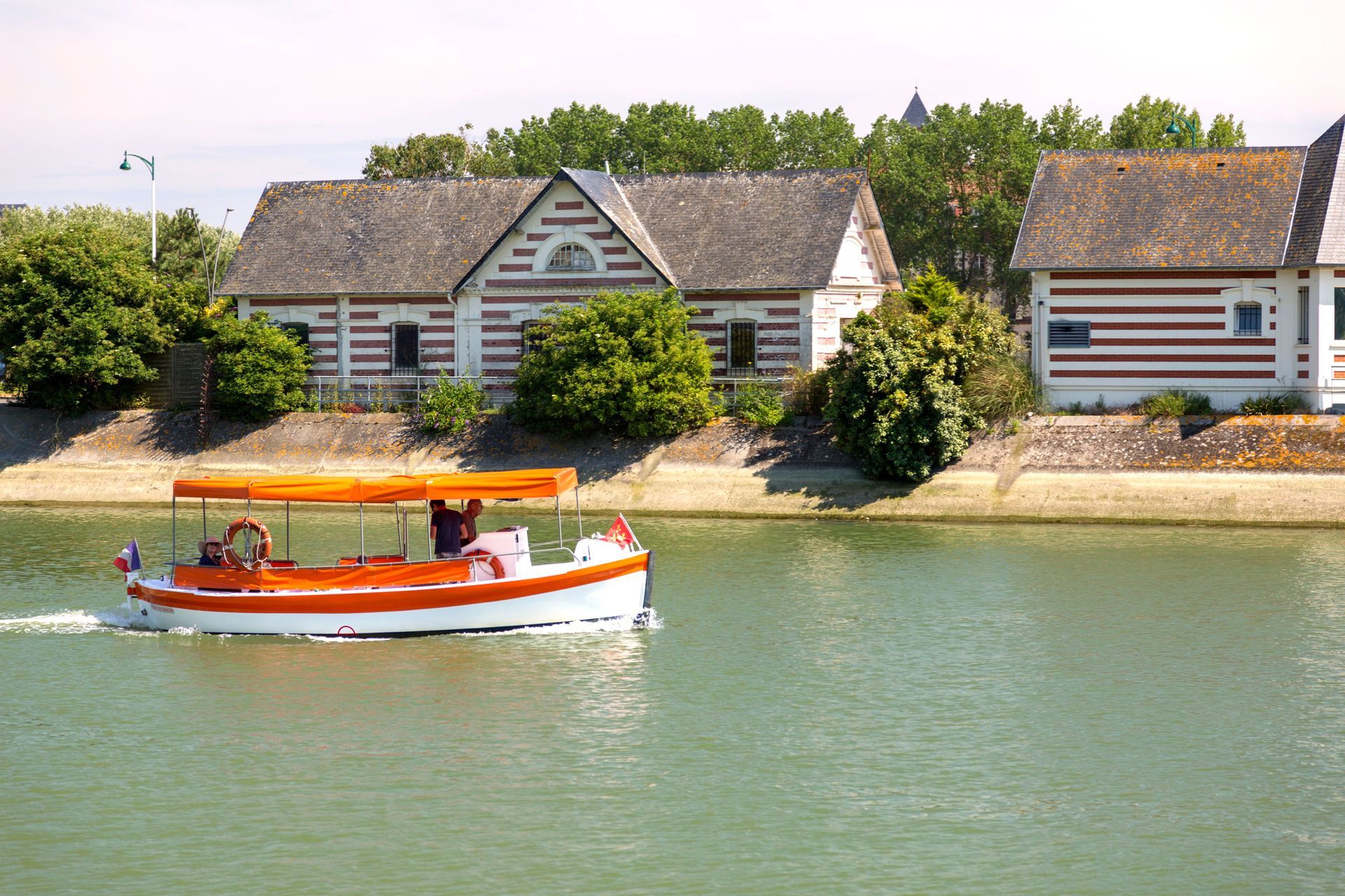 Small orange boat on a calm river in front of striped riverside cottages and green trees