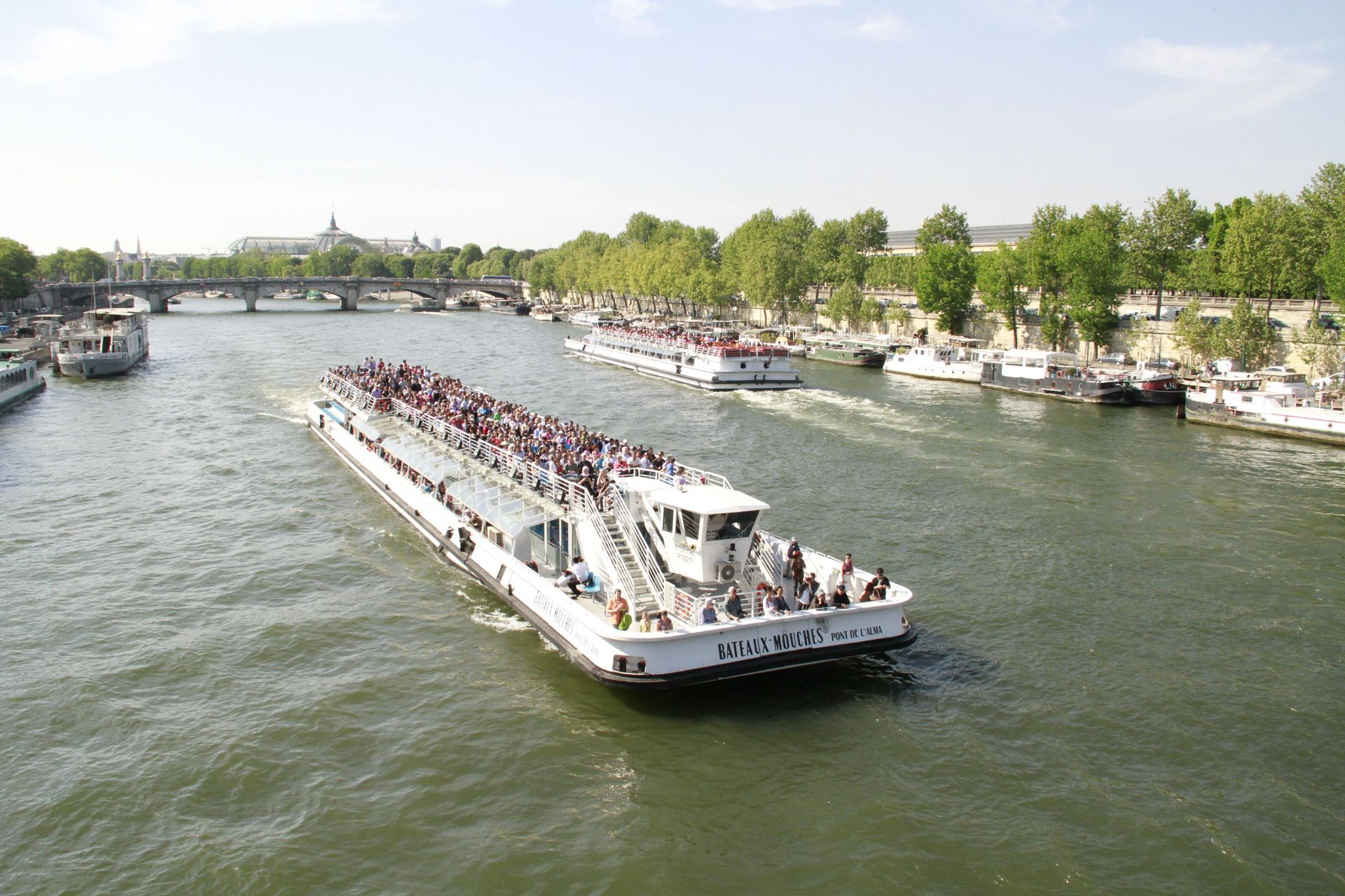 Long white passenger boat full of people cruising a wide river past a green waterfront promenade