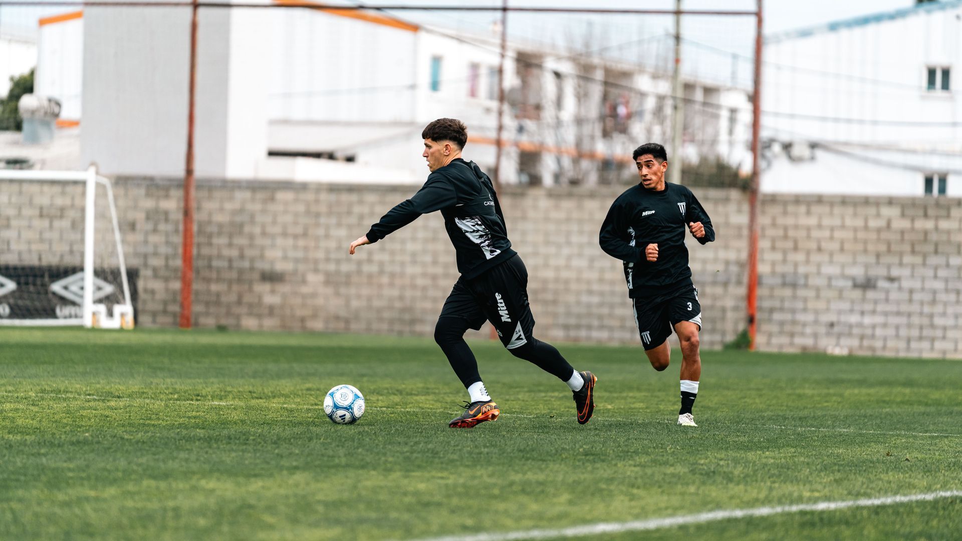 Two soccer players in black training gear on a field, one dribbling a blue-and-white ball