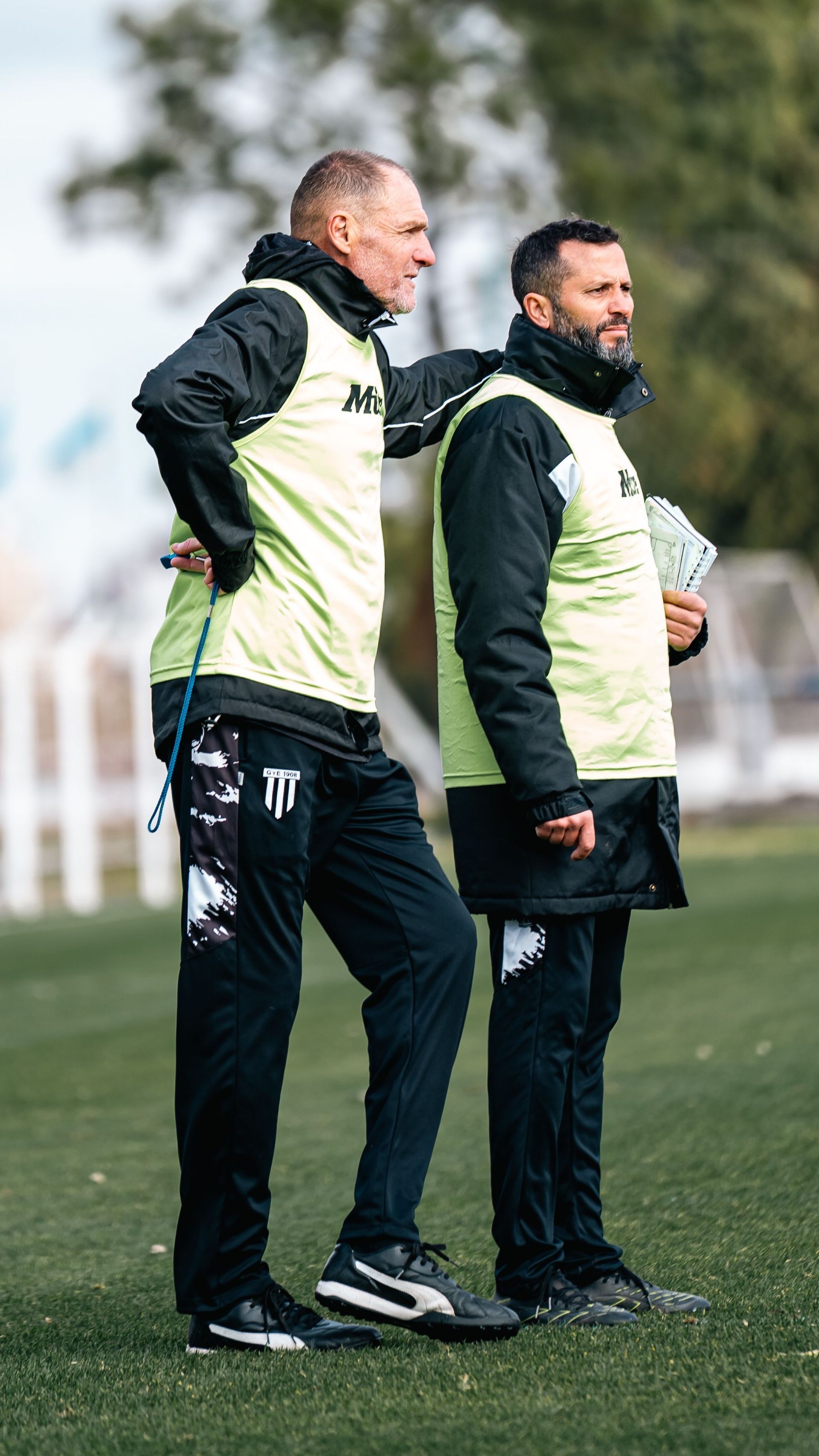 Two men in black and cream sportswear standing on a grassy field during training