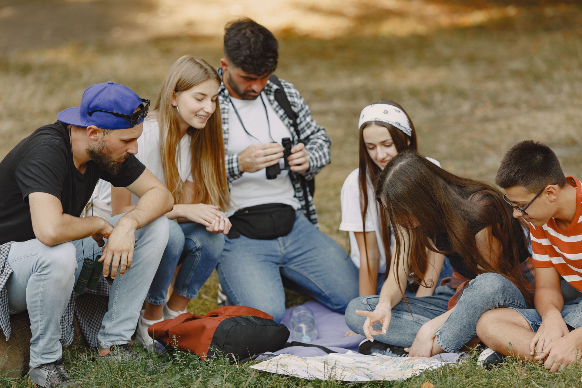 Group of friends sitting on grass outdoors, gathered around a map and backpack on a sunny day