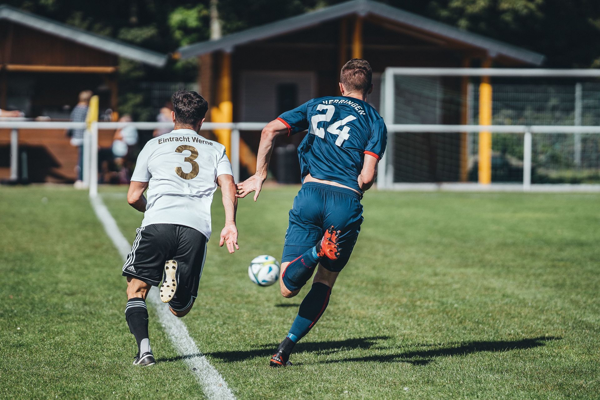 Two soccer players sprint after the ball on a grassy field, one in white and one in blue, near the goal line