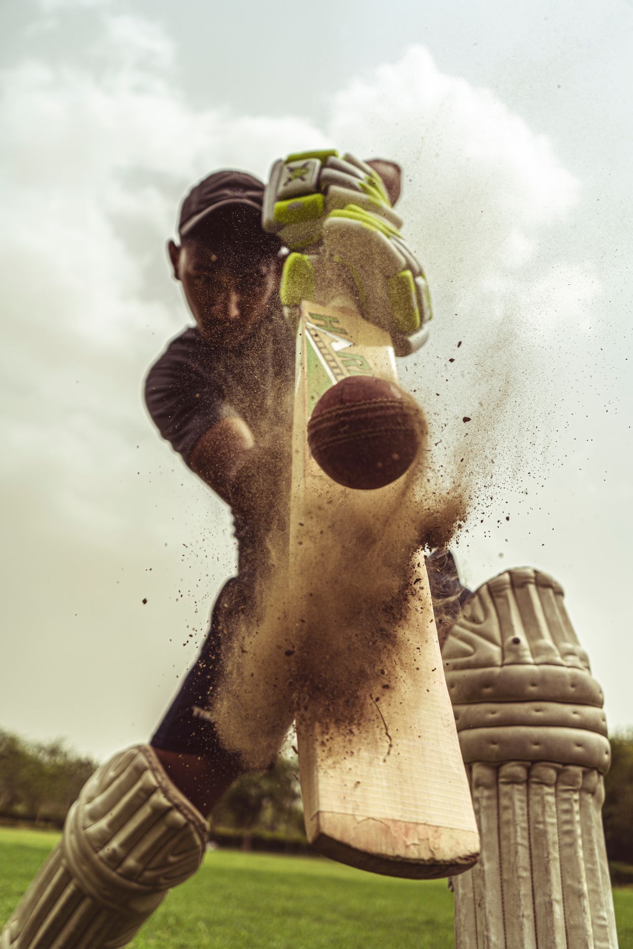 Cricketer swings bat as dust bursts up during a powerful hit on a sunny field