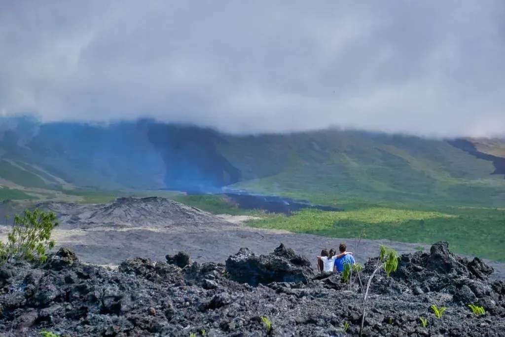 People standing on a rocky volcanic landscape with smoke rising from a crater under cloudy skies