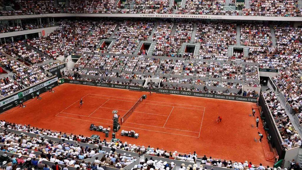 Wide view of a clay tennis court with a match in progress and packed stadium seating