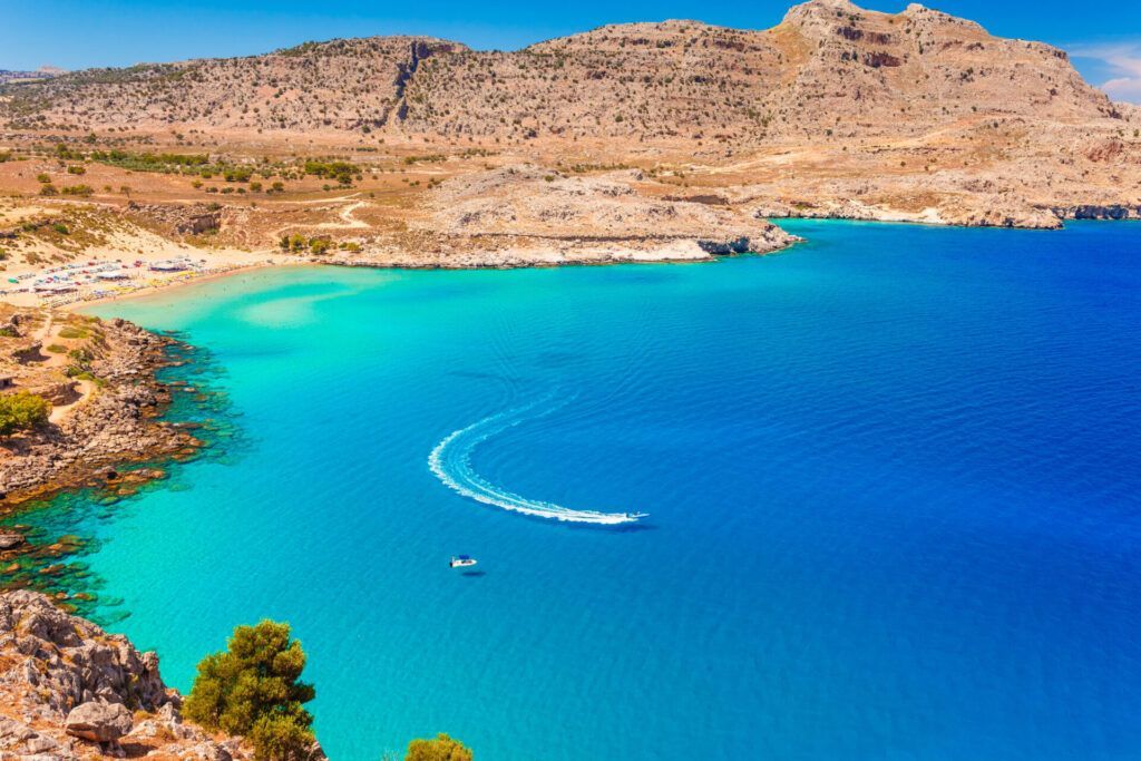 Turquoise bay with a small speedboat leaving a white wake beside rocky desert cliffs
