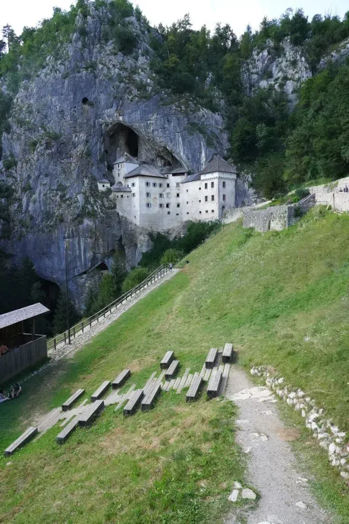 Stone monastery built into a cliff above a grassy hillside, with mountains and trees in the background