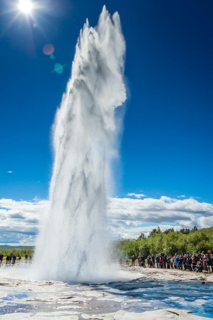 Tall white geyser erupting against a blue sky with a crowd watching nearby