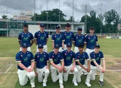 Cricket team posing on a field, wearing navy jerseys and white pants, with a cloudy sky behind them