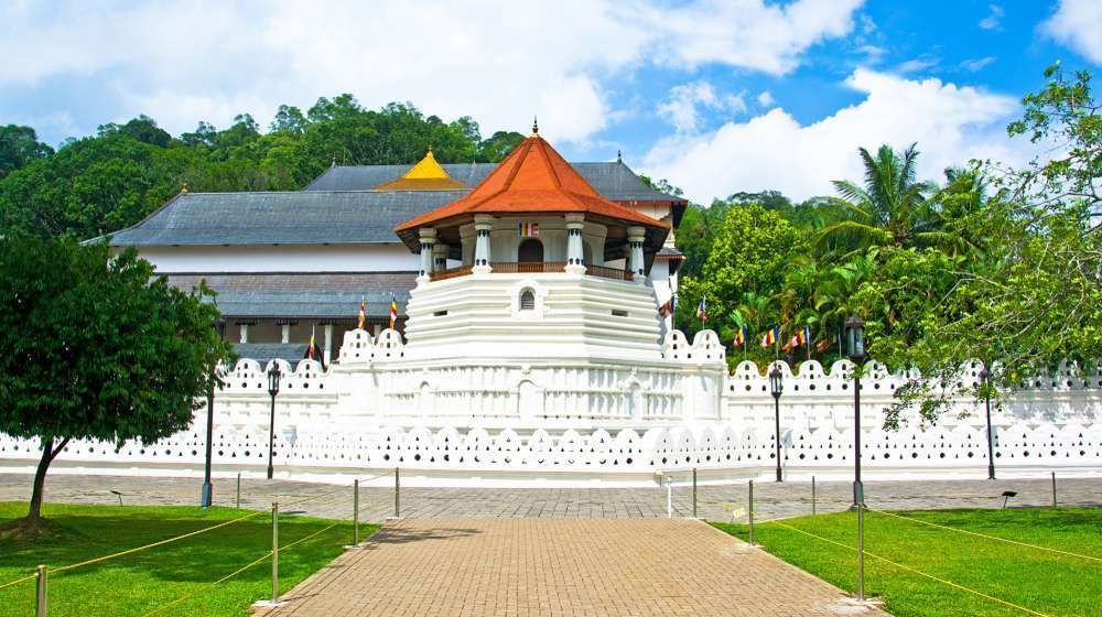 White Buddhist temple with orange roof, white fence, and green trees under a bright blue sky.
