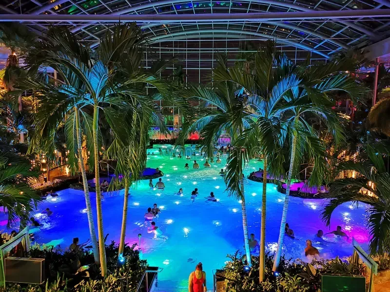 Indoor tropical waterpark with glowing blue and purple pools, palm trees, and swimmers under a domed ceiling