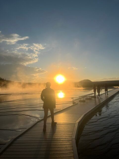 Person silhouetted on a boardwalk at sunrise over calm water, with another figure in the distance.
