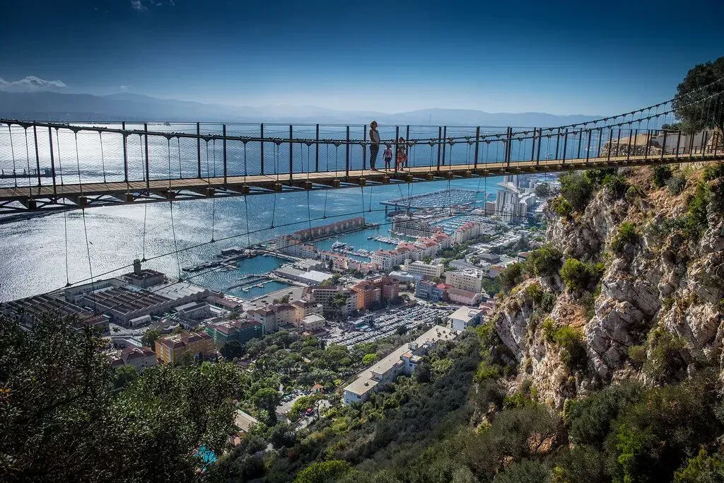 Suspension bridge over a coastal town and bay, with hills and a bright blue sky.