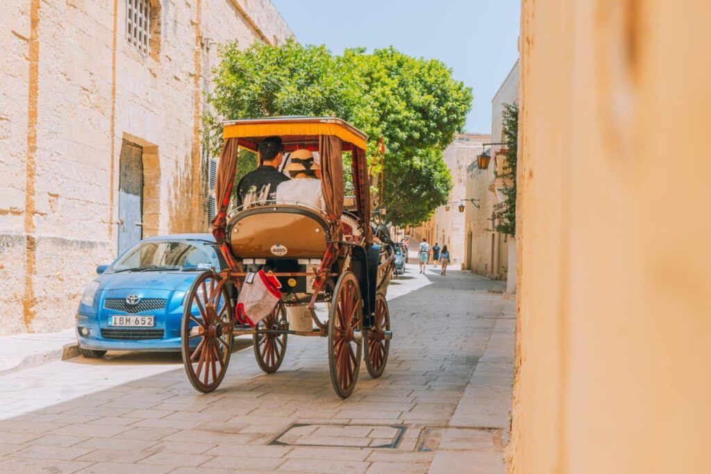 Horse-drawn carriage on a narrow sunlit street beside a blue car and peach-colored wall
