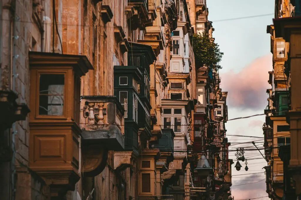 Narrow city street lined with old apartment balconies at sunset, with cables overhead and pink clouds in the sky