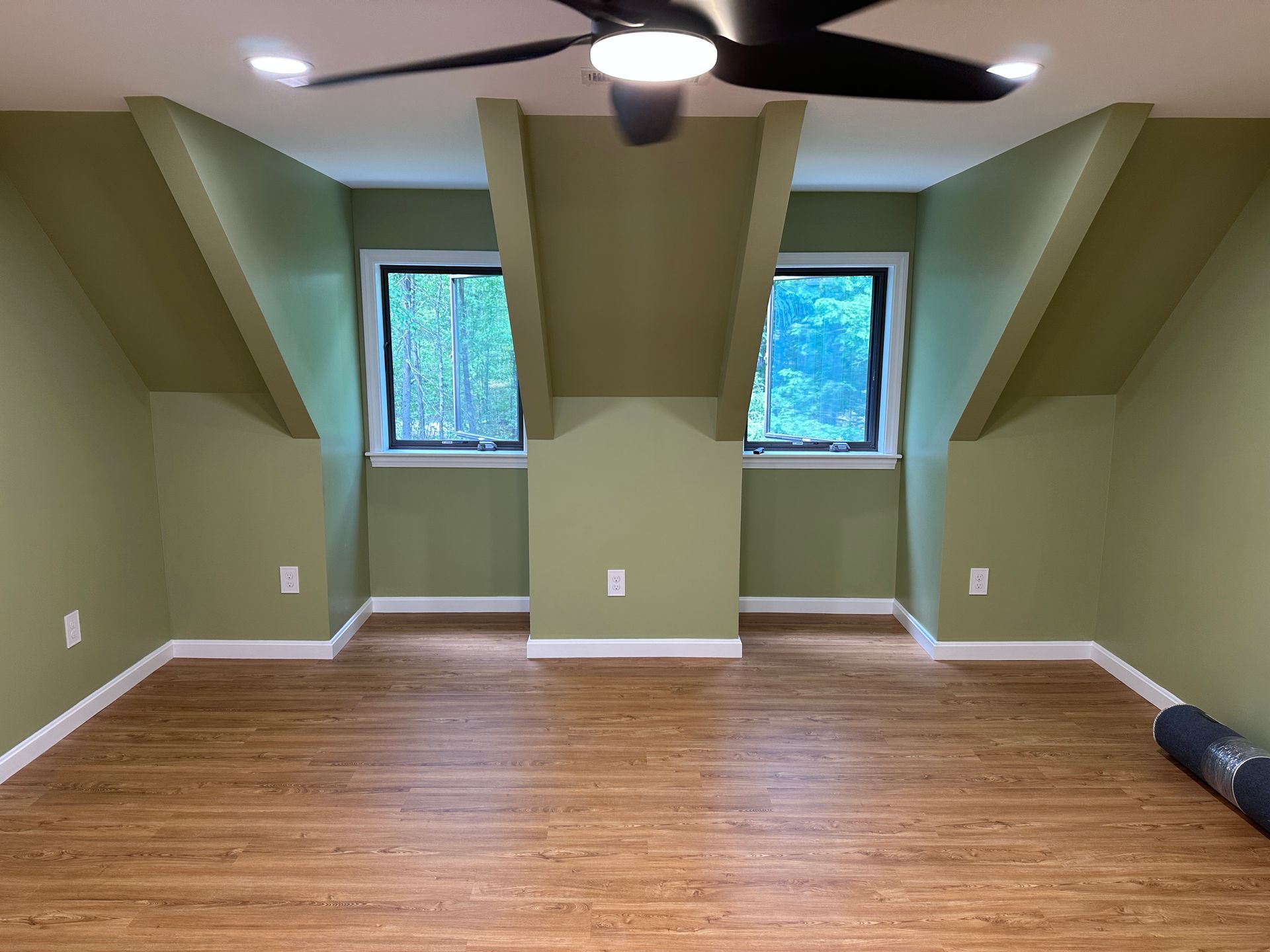 Empty green attic room with wood floor, two windows, and a ceiling fan center.