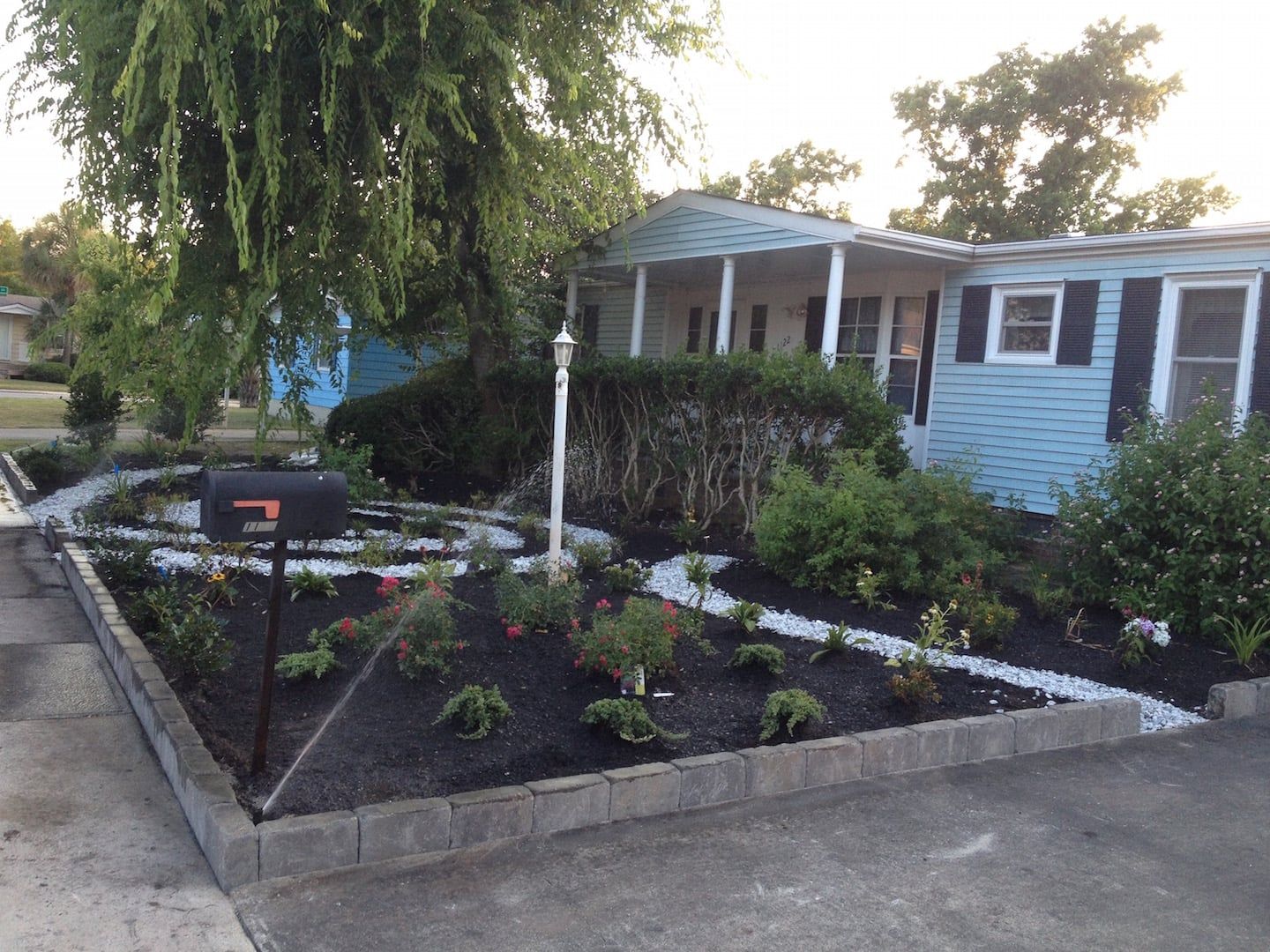 Front yard garden with flowers, a mailbox, and a light blue house behind a stone border.