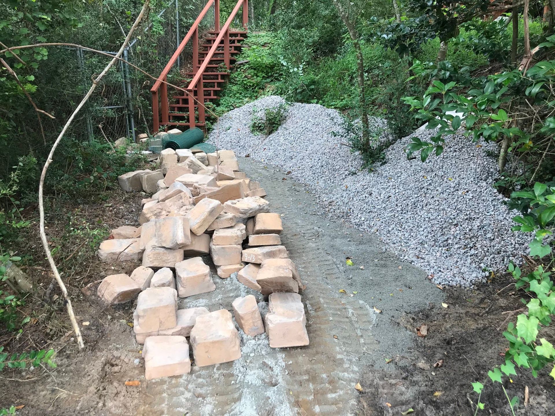 A pile of bricks and gravel beside a forest path with a red staircase in the background