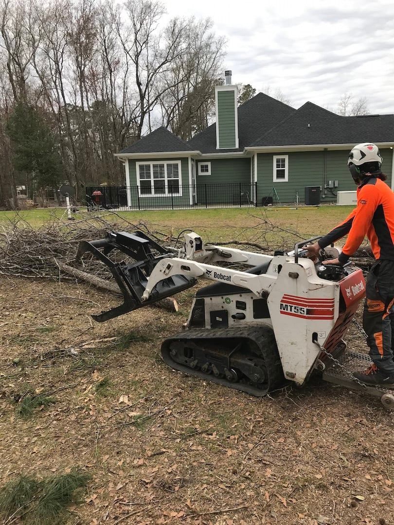 Worker operating a skid steer to clear brush in a yard beside a green house