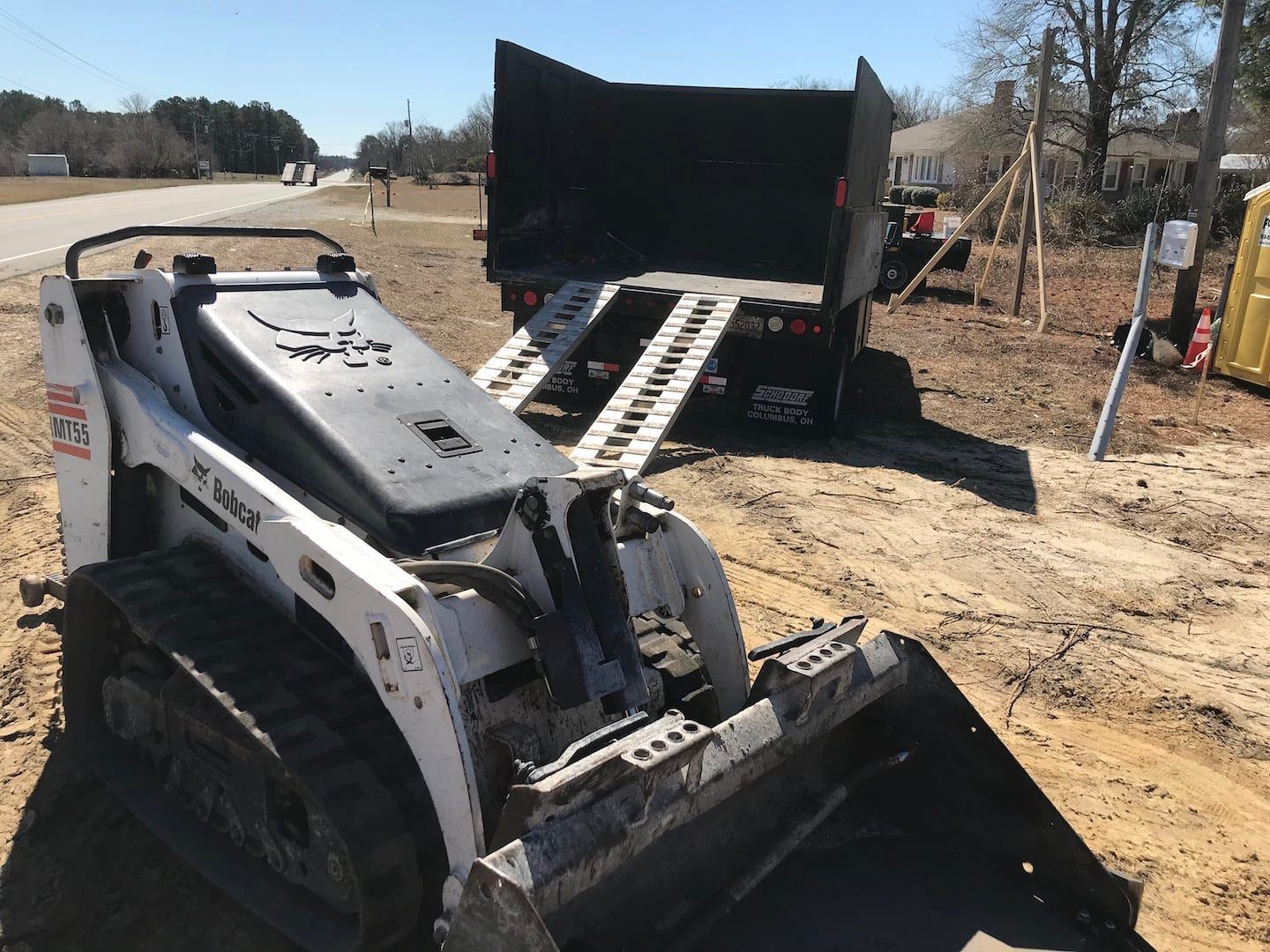 White skid-steer loader loading dirt into a black dump truck on a roadside construction site
