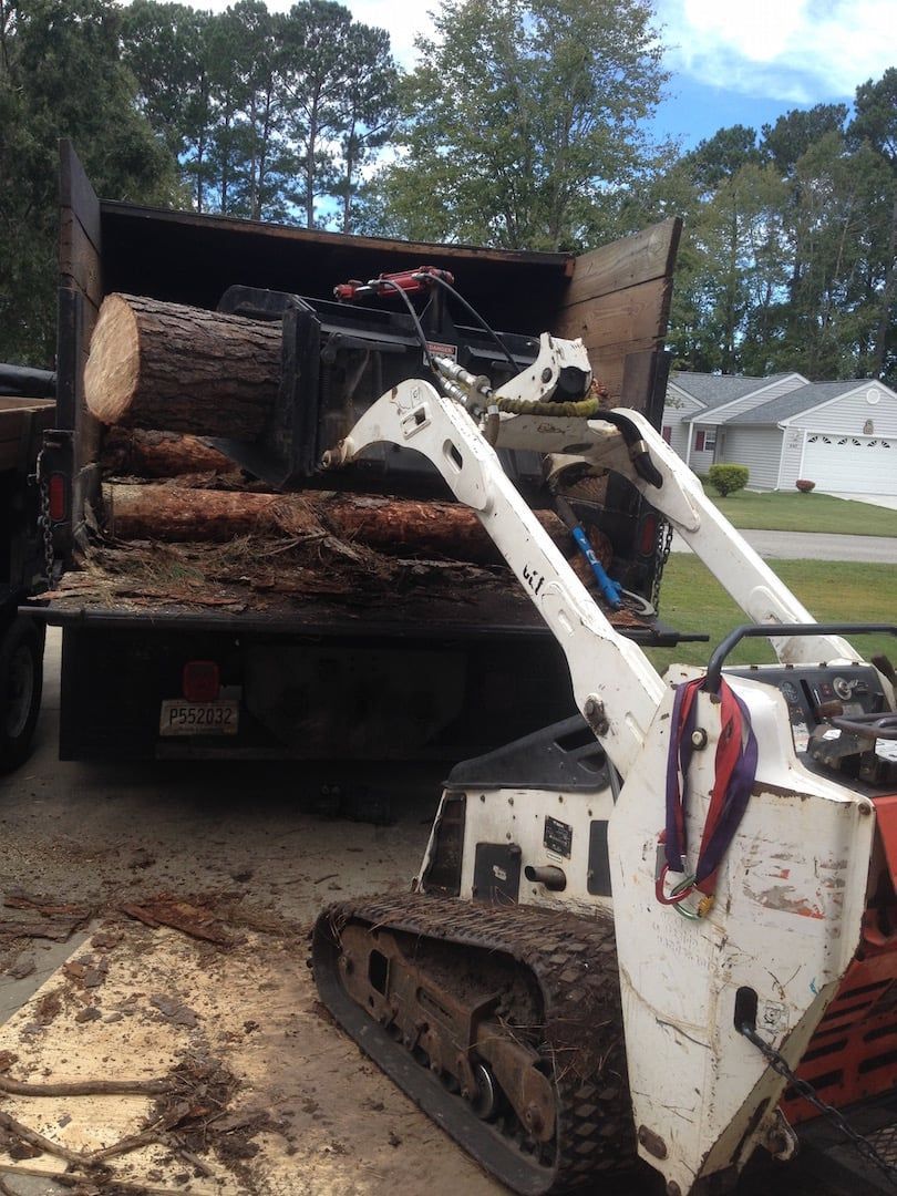 Small skid-steer loader unloading logs from a truck bed outdoors.