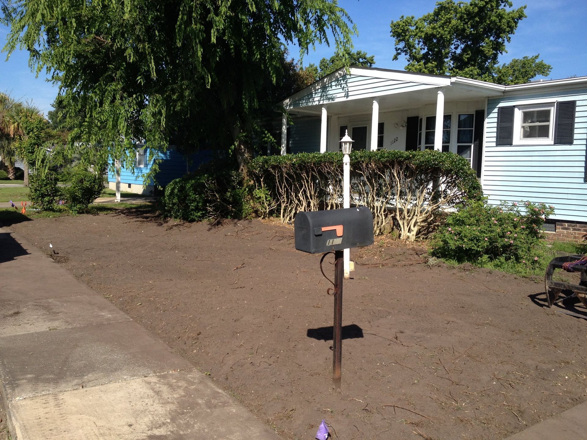 Black mailbox in front of a light blue house with a porch and bare yard, shaded by a tree