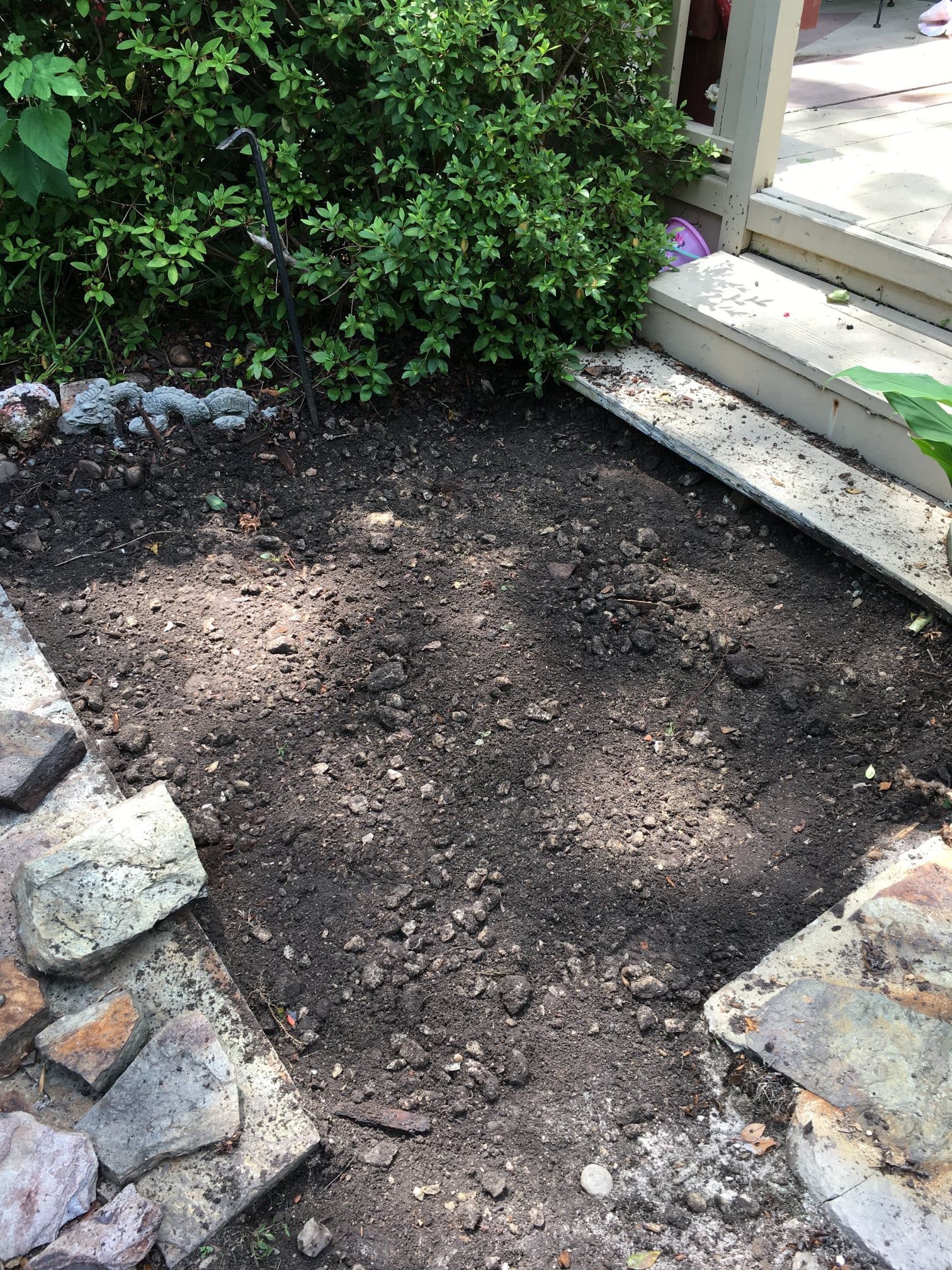 Bare garden patch bordered by stones, beside green shrubs and a house step
