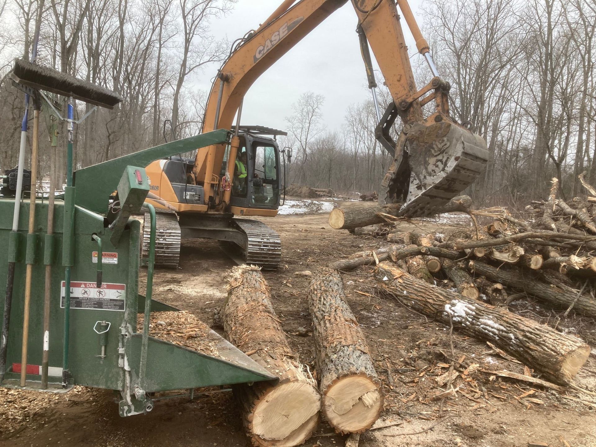 Excavator loading cut logs beside a green wood chipper in a wooded clearing