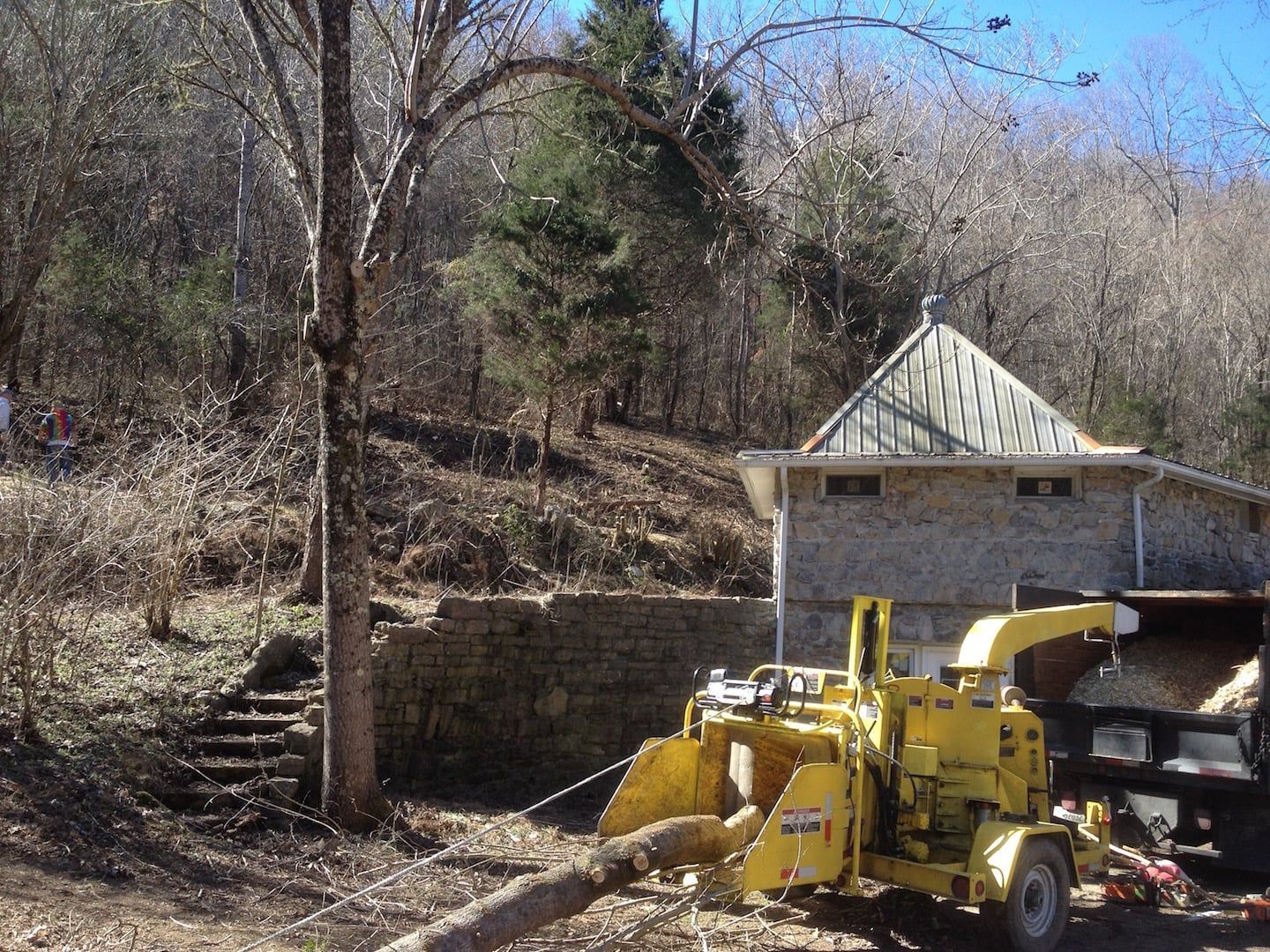Yellow backhoe parked beside a stone house on a wooded hillside in winter