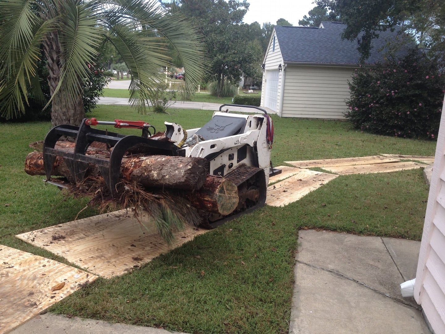 Skid steer carrying a large tree trunk on a driveway beside a lawn and house