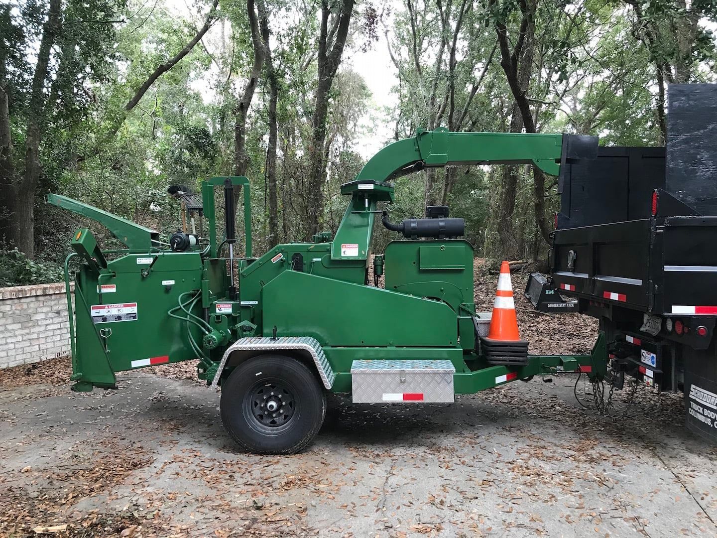 Green wood chipper attached to a truck in a wooded area, with an orange traffic cone nearby.
