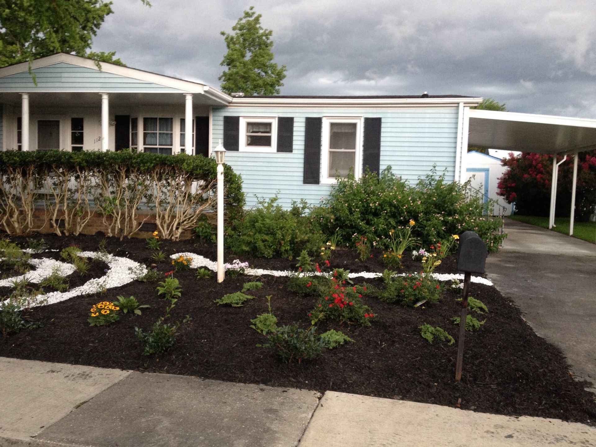 Landscaped front yard with flower beds, a light blue house, and a driveway under cloudy skies