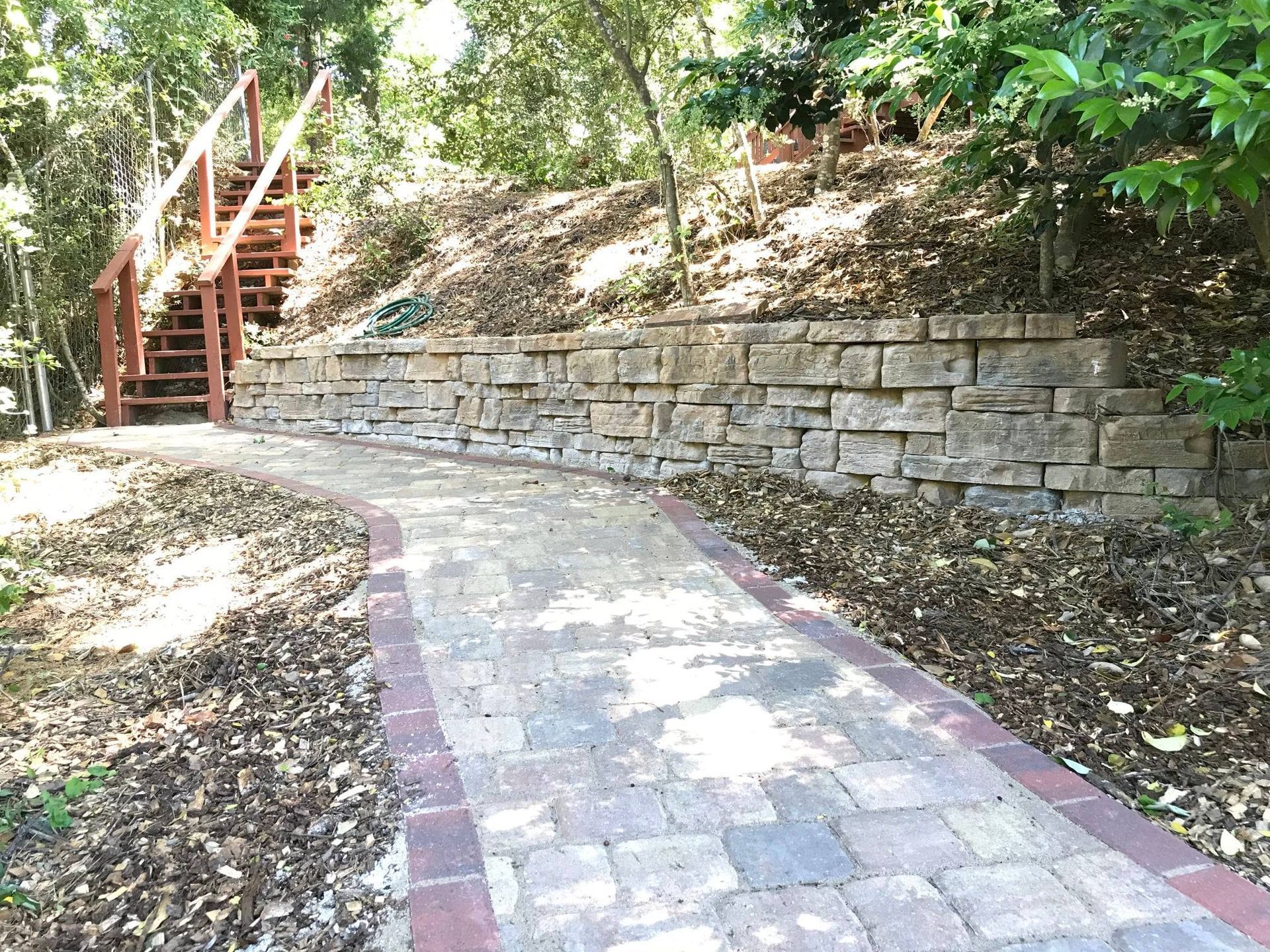 Curved stone walkway beside a retaining wall, leading to wooden steps in a wooded area.