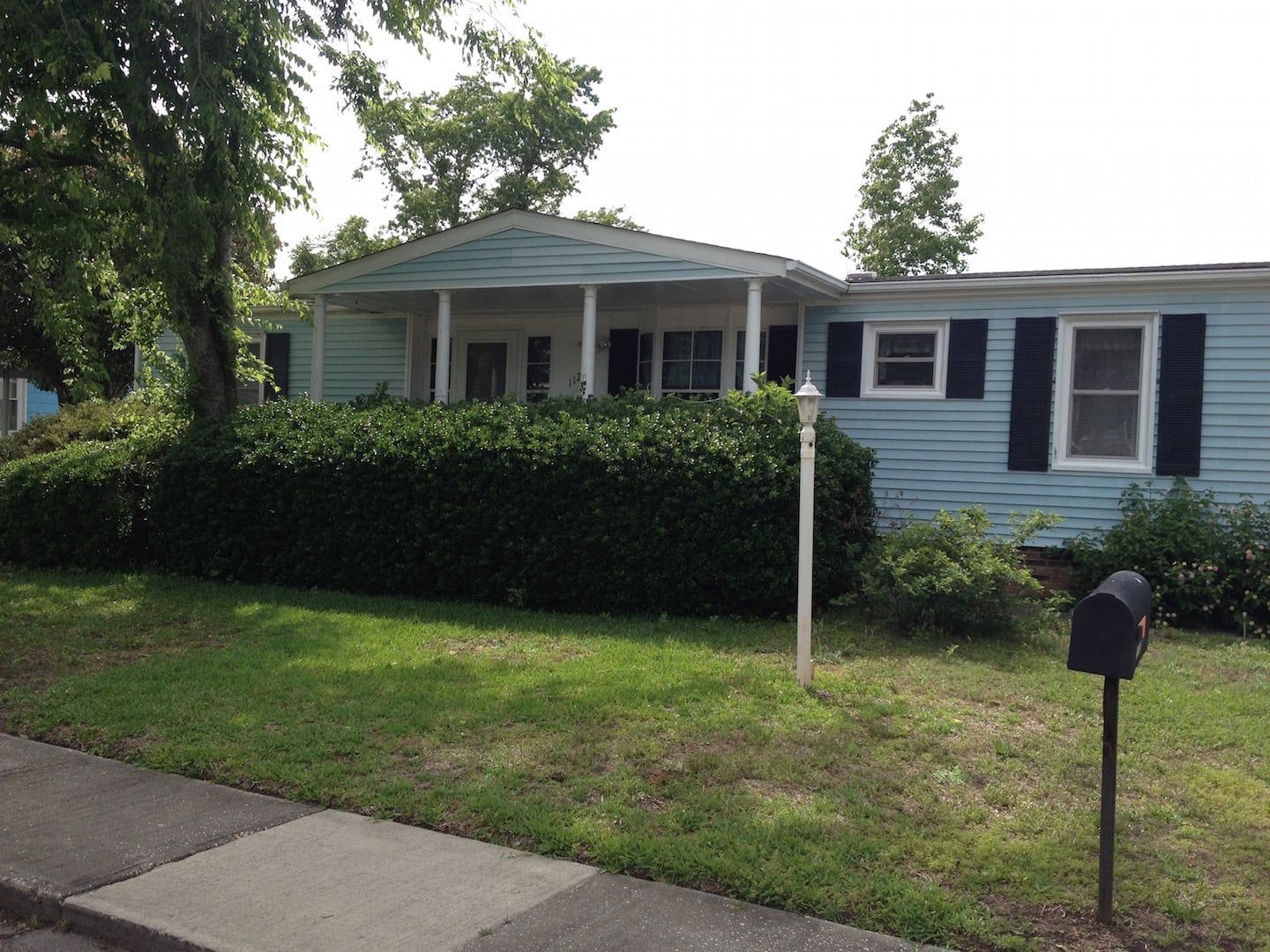 Blue house with white porch behind hedge and lawn, viewed from the sidewalk
