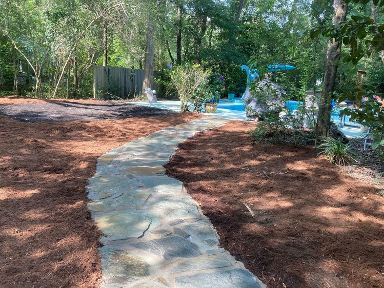 Curving stone garden path through mulched woodland with trees and a small blue fountain.