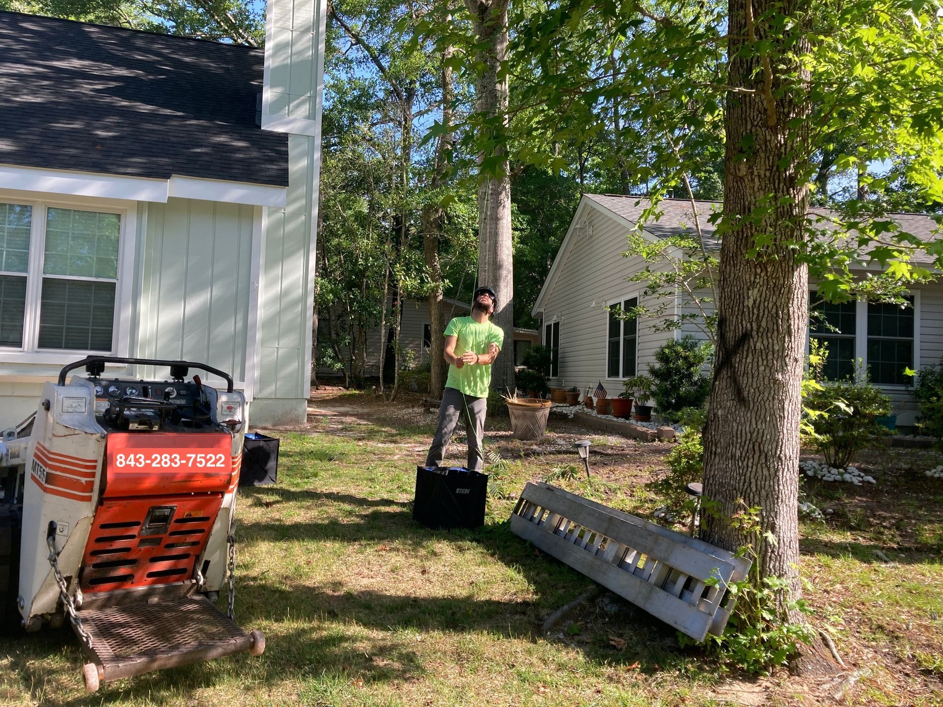 Worker on lawn mower in front yard beside a house, with a red dumpster and trees nearby.