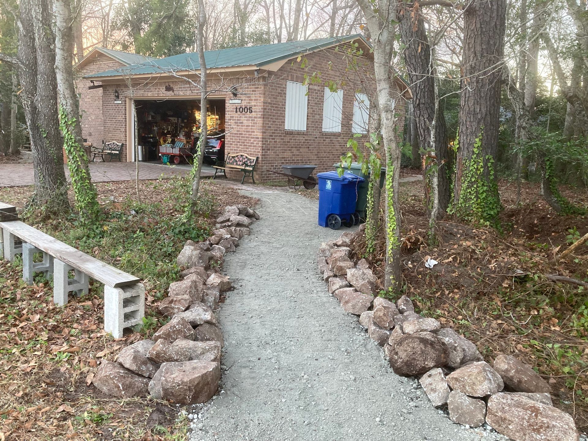 Gravel path leading to a brick shed in a wooded yard, with blue trash bins and a white bench nearby