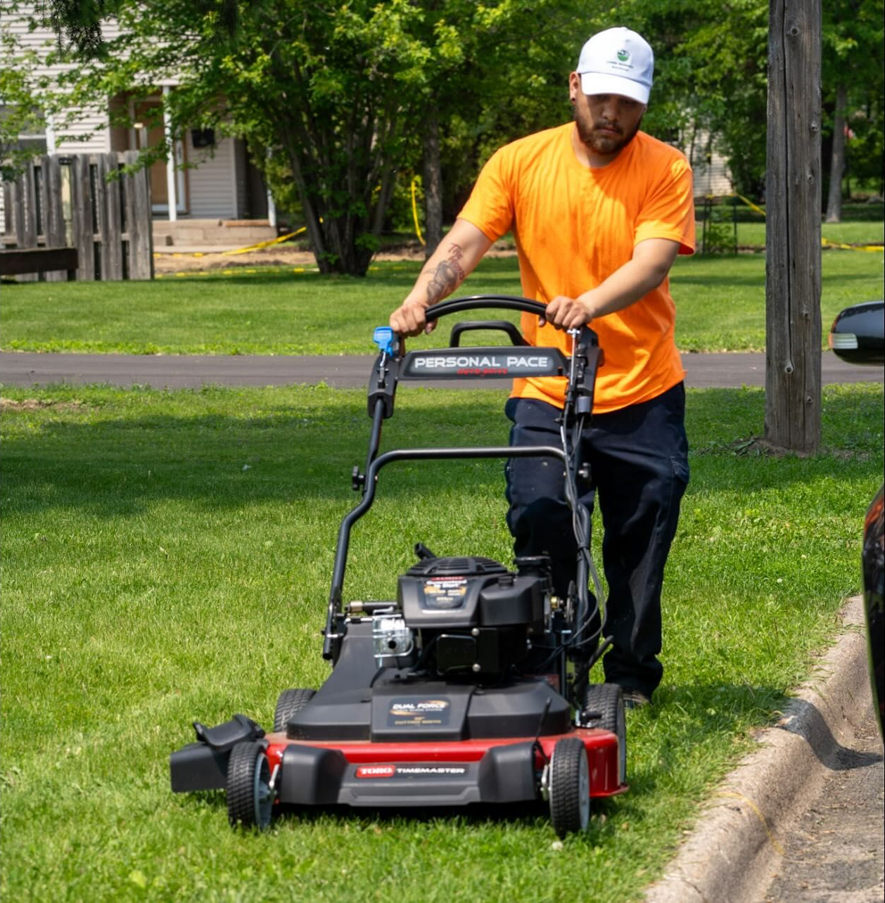 Worker in orange shirt pushing a red-and-black lawn mower across a grassy yard