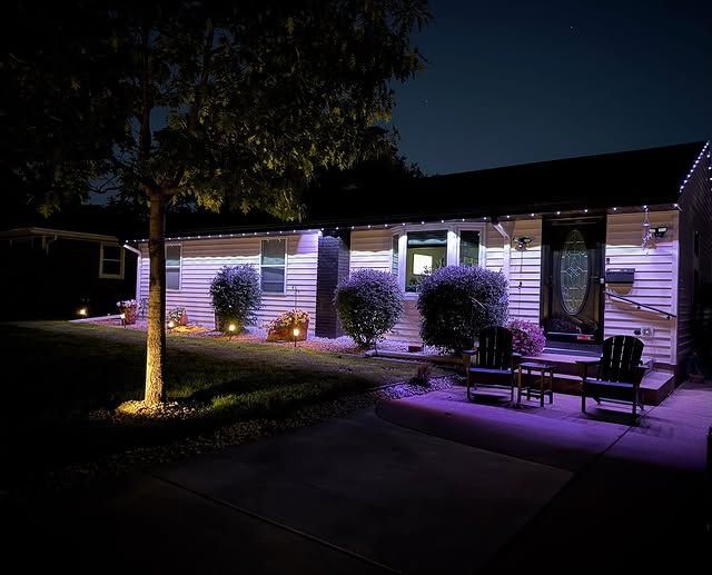 Purple-lit house exterior at night with trees, bushes, and a porch seating area