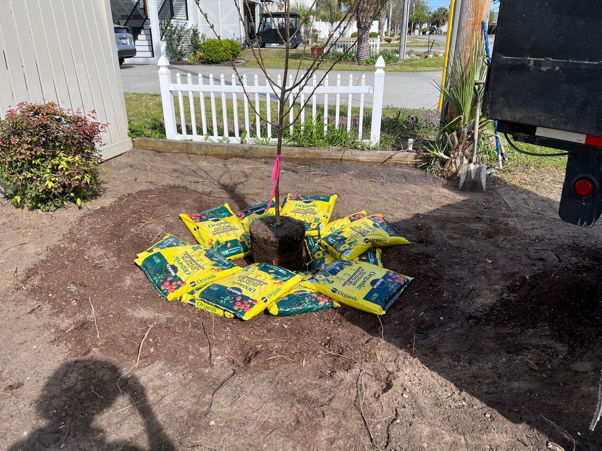 Freshly planted garden bed with a young tree, surrounded by yellow-and-green mulch bags and a white fence.