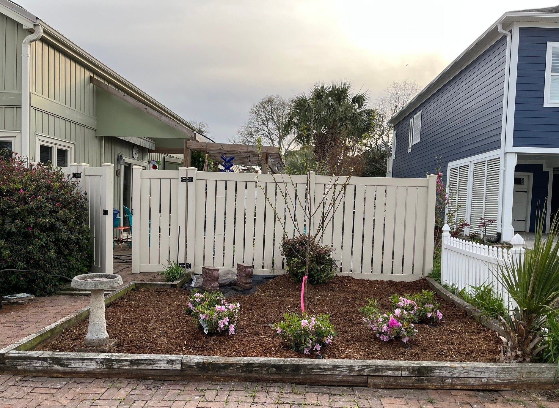 Small garden bed with flowers in a backyard between two houses, bordered by a white fence.