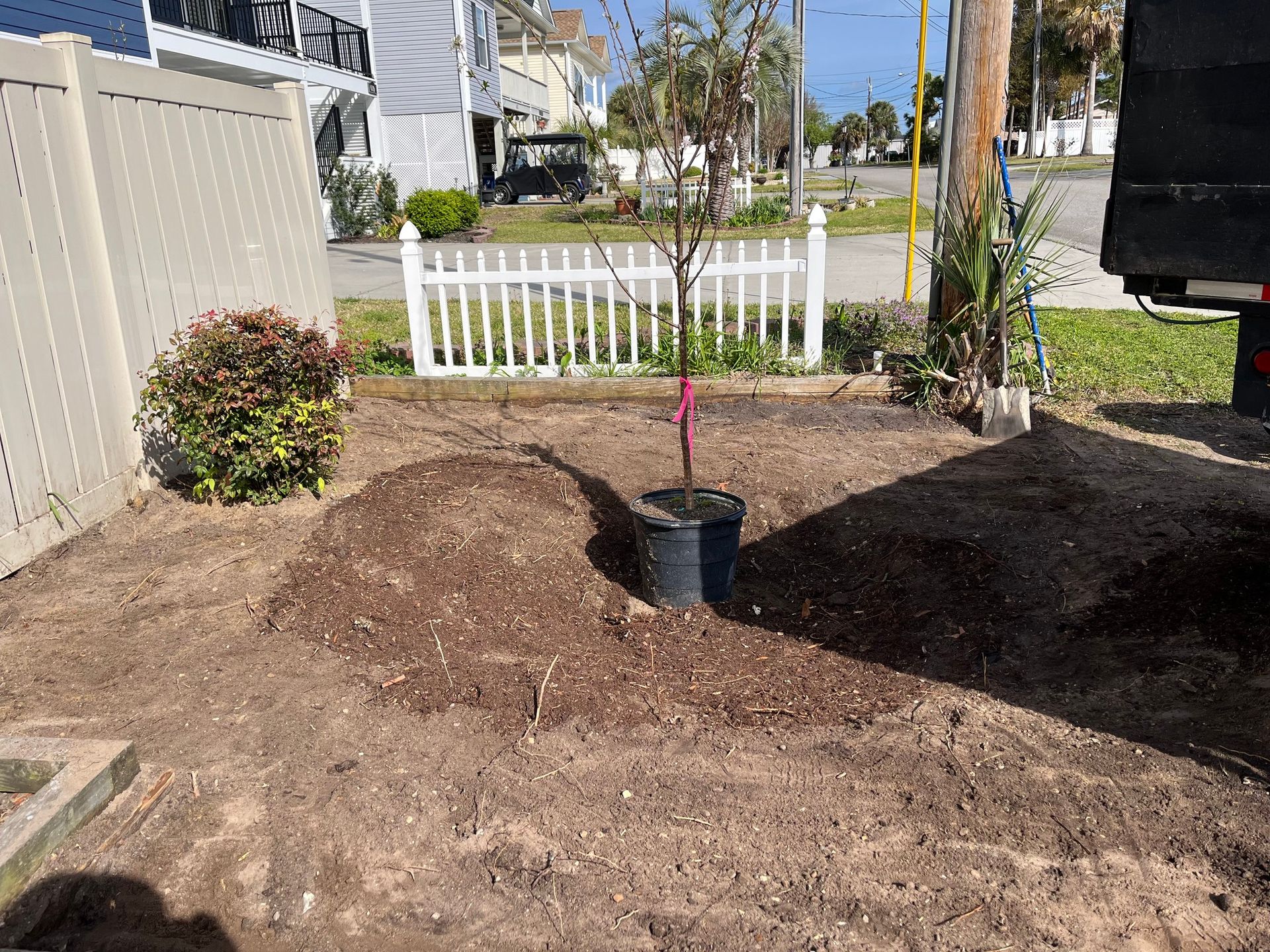 Freshly dug yard bed with mulch, a black bucket, and a pink marker near a white fence