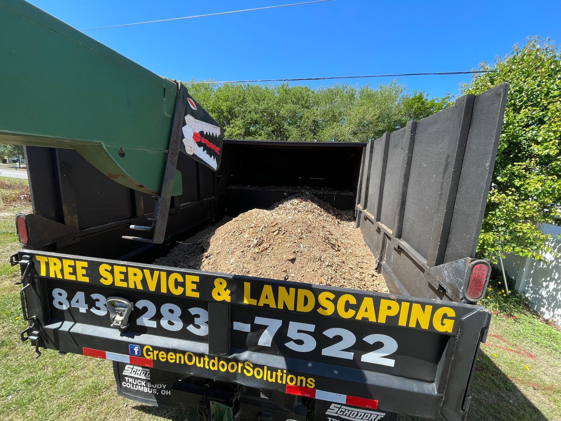 Tree service truck unloading wood chips into a trailer at a sunny job site