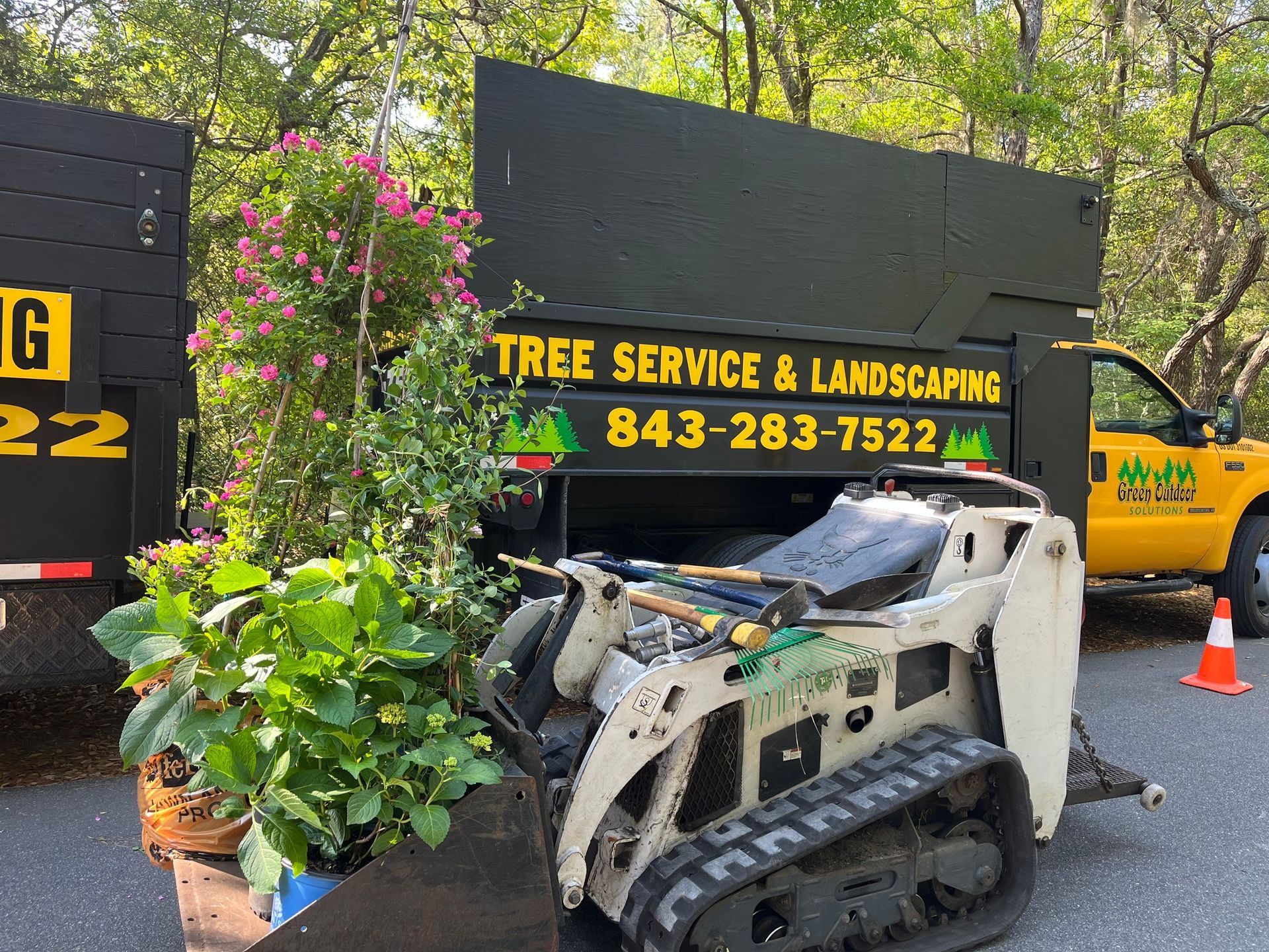 Compact track loader beside tree service truck and potted plants in a landscaping yard