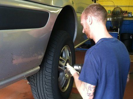 Mechanic using a power tool to remove a tire from a silver car in a garage.
