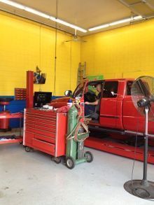 A red truck in a yellow-walled auto shop, a person in the driver's seat, near a tool cart and welding gear.