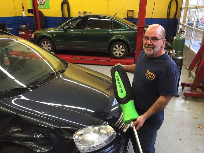 Man with a Snap-on tool smiles next to a black car in a garage; a green car is behind him.