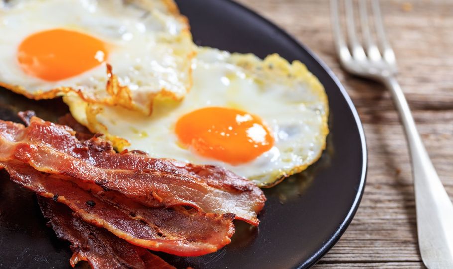 Two fried eggs with orange yolks, crispy bacon, on a black plate with a fork on a wooden table.