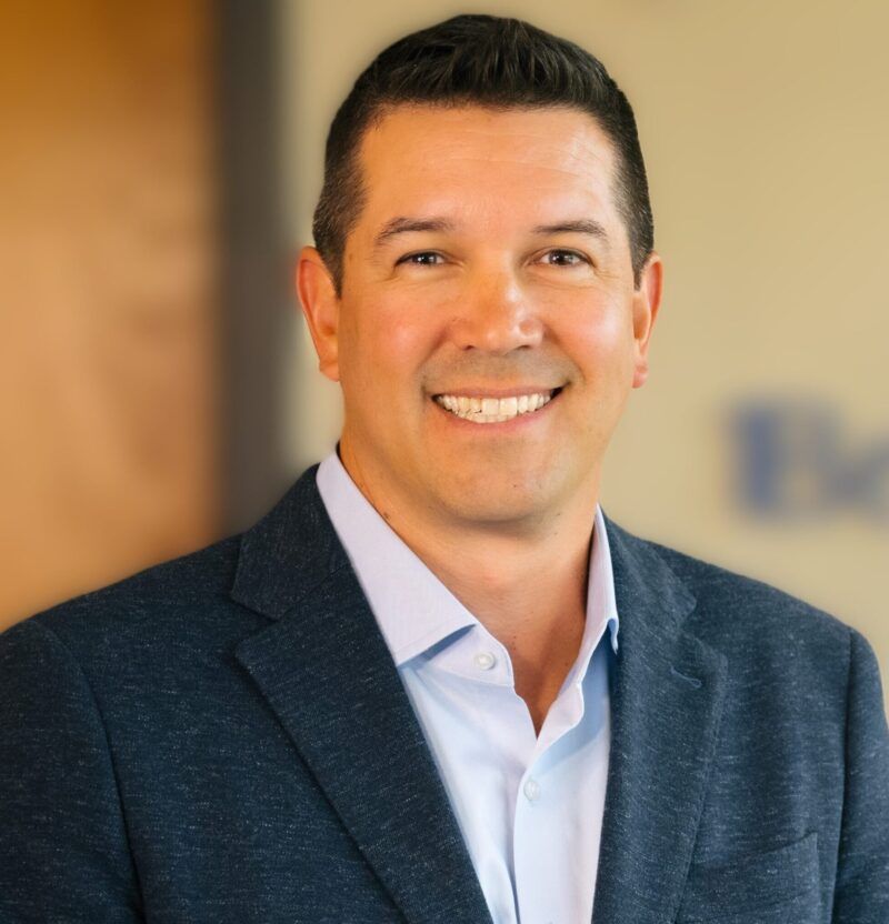 Smiling man in a navy blazer and light blue shirt against a blurred office background