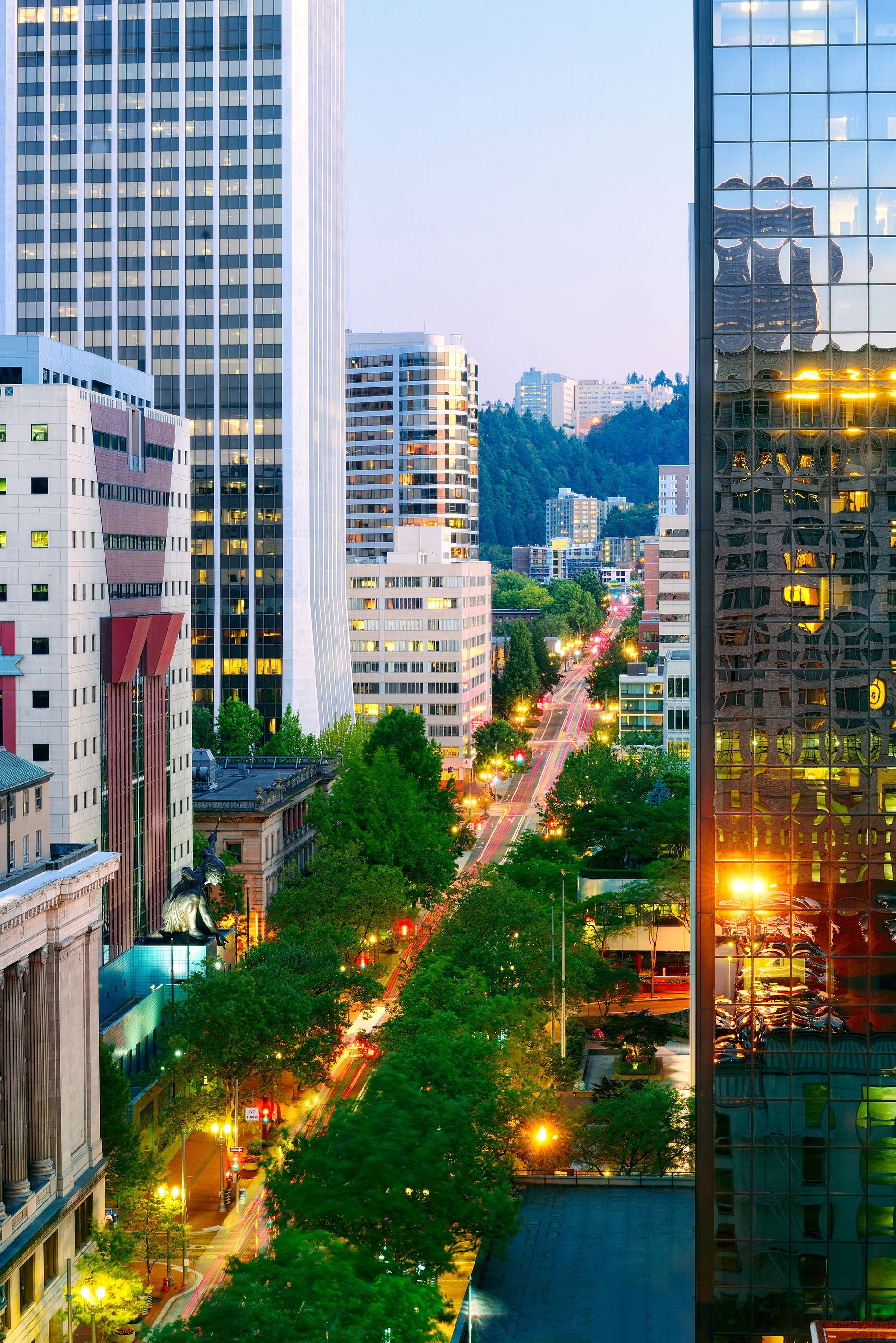 City street lined with trees between tall office towers at dusk, with lights reflecting in glass buildings